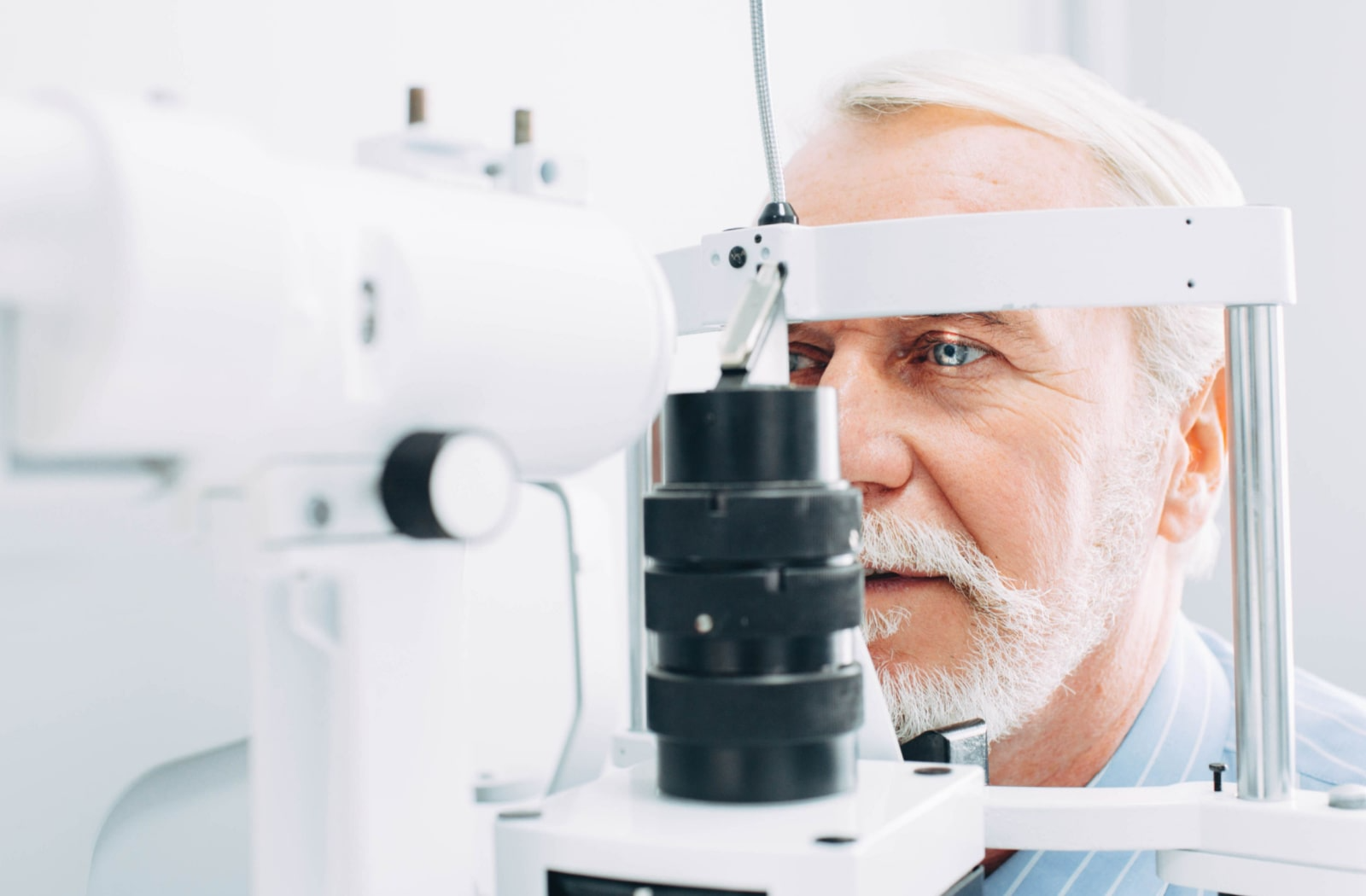 An older man sitting and placing his face on optometry equipment so the optometrist can examine his eyes for diseases, like diabetes.