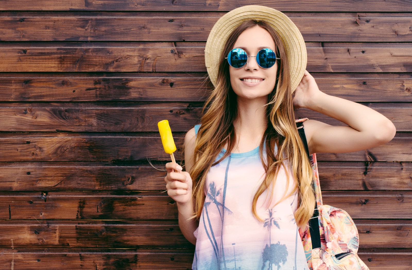 A woman standing against a wood panel background, holding onto a popsicle stick. She's holding onto her sun hat with her other hand, and wearing polarized sunglasses.