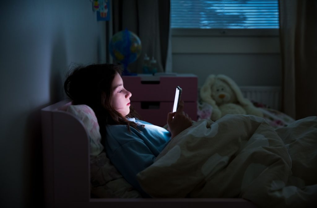 A young child lying in bed scrolling on her phone in the dark.