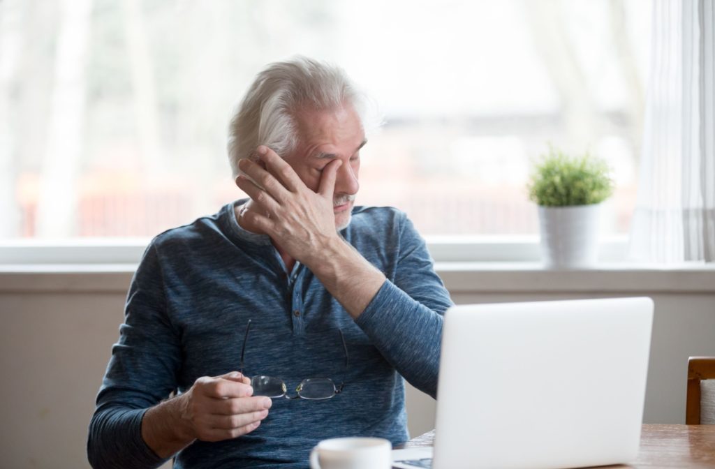 An adult taking off their glasses while working at home to rub their eyes in frustration due to wintertime dry eyes.