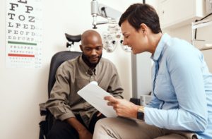 A smiling optometrist holding a clipboard and showing a patient how to read the numbers on their prescription after an eye exam.