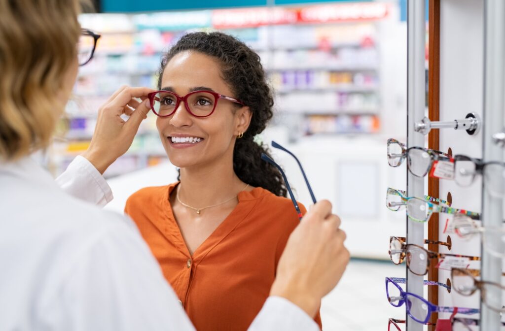 Smiling person trying on red eyeglasses with the help of an optician in a store.