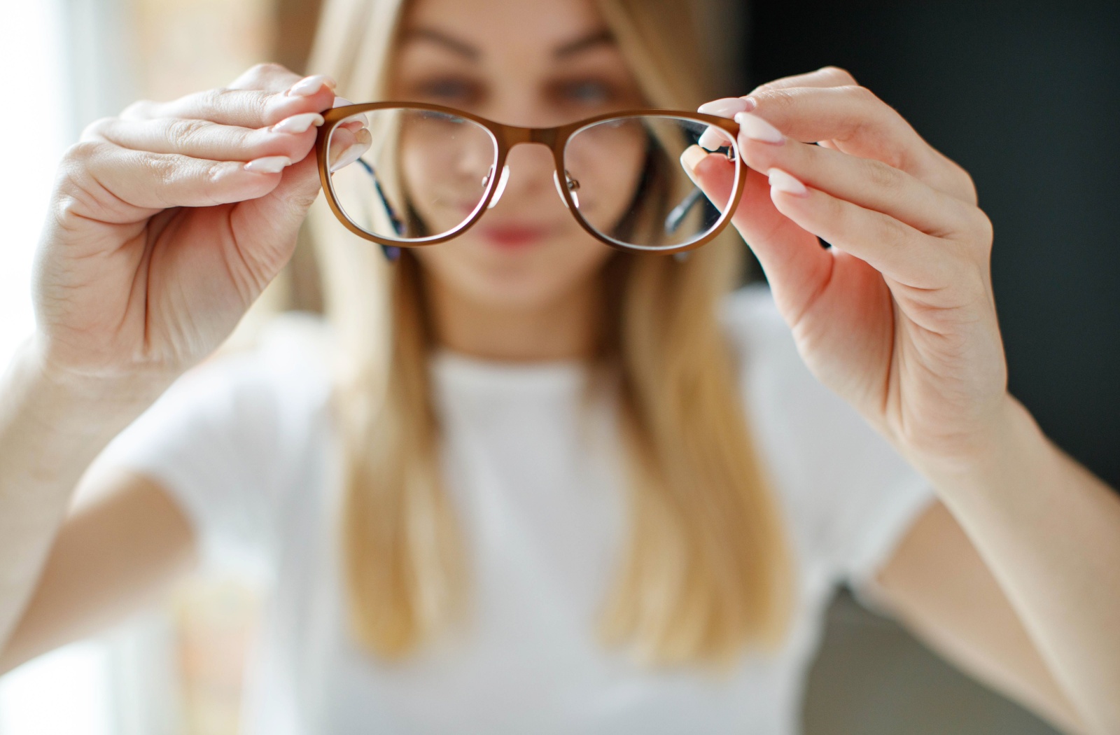Blurry image of a woman with long blonde hair holding up glasses, which are clear and focused in the foreground.