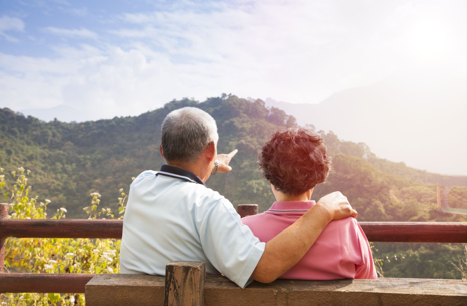 A senior couple sits on a bench looking into the distance at green mountains
